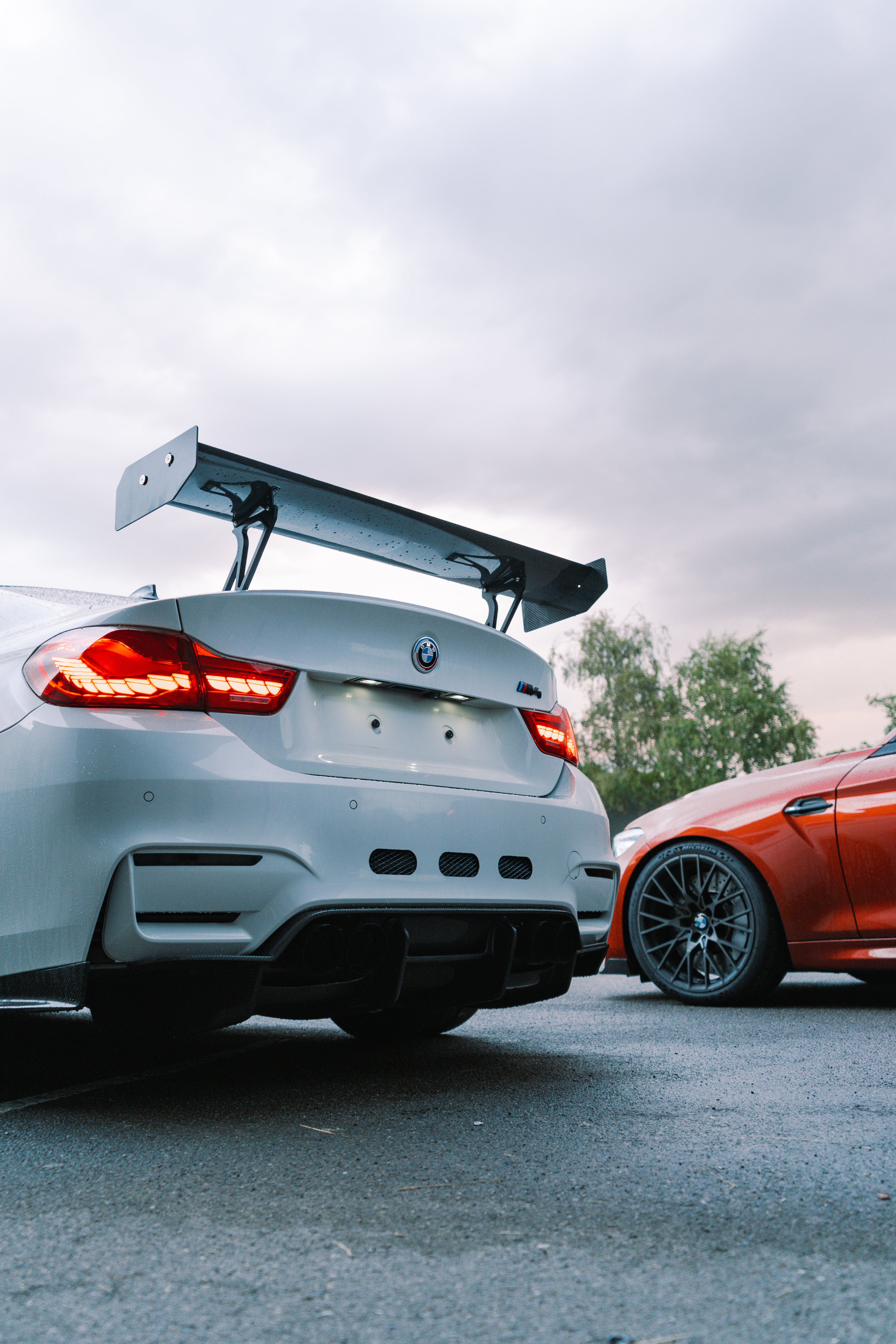 Two cars, one white and one orange, parked side by side on a road with a cloudy sky.